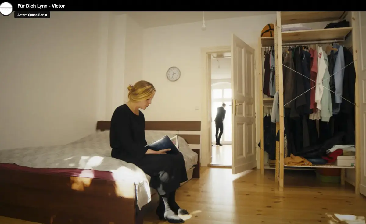 Eine Frau sitzt auf einem Bett in einem schlichten Schlafzimmer und hält ein Buch im Schoß. Im Hintergrund steht ein Mann im Türrahmen des angrenzenden Raums. Rechts ist ein offener Kleiderschrank zu sehen.