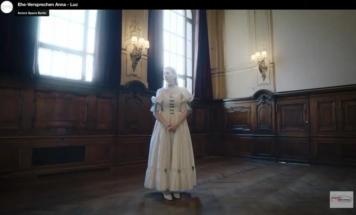 Eine Frau in einem weißen historischen Kleid steht in einem großen Saal mit dunkler Holzvertäfelung und hohen Fenstern. Warmes Licht fällt rechts aus dem Fenster in den Raum.