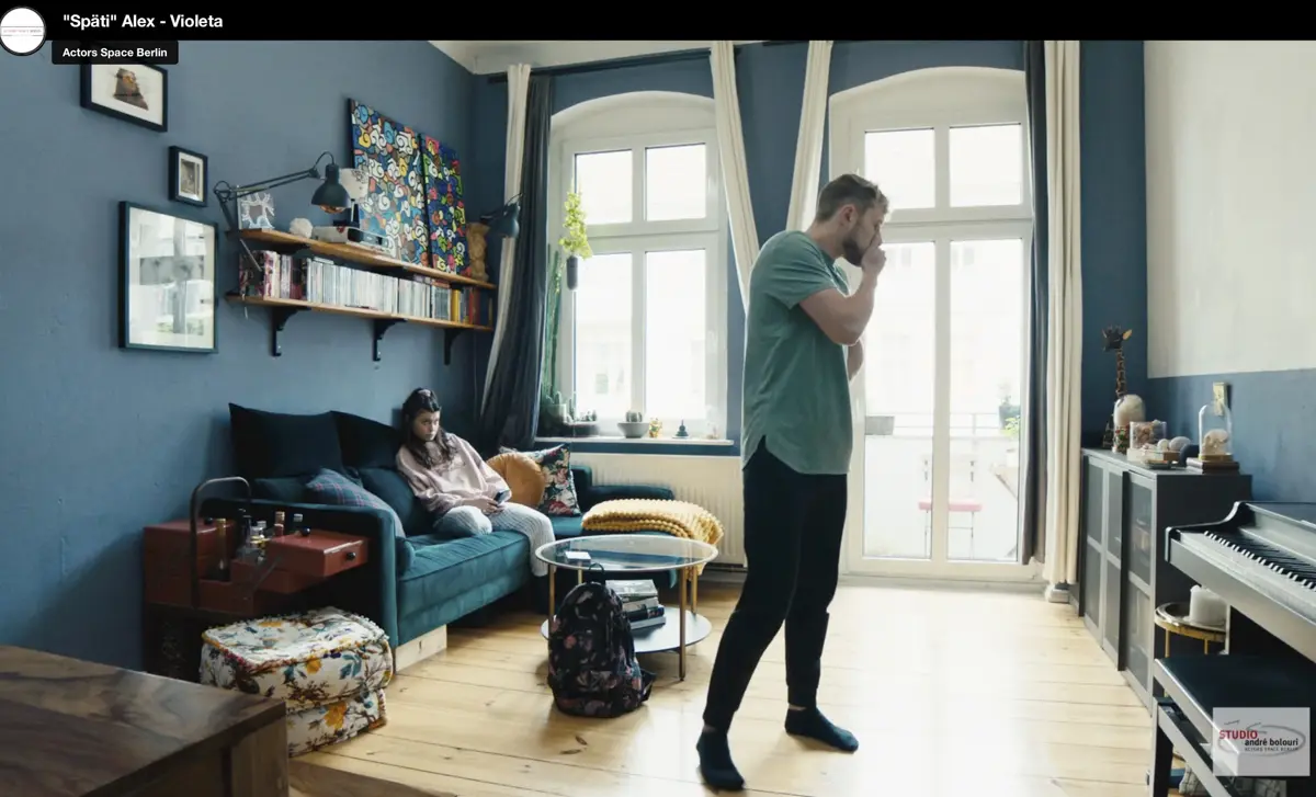 Ein Mann steht in einem hellen Wohnzimmer vor großen Fenstern und hält sich nachdenklich das Gesicht, während eine junge Frau auf einem Sofa sitzt und auf ihr Handy schaut. Der Raum ist modern eingerichtet mit blauen Wänden und vielen Dekorationen.