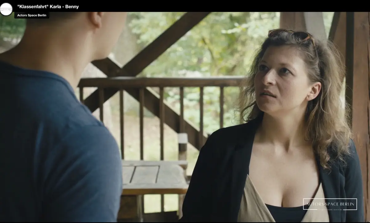 Eine Frau im blauen historischen Kleid steht in einem reich verzierten Raum mit Kronleuchter, hohen roten Vorhängen und antiken Möbeln. Der Raum wirkt wie ein Salon oder ein historisches Esszimmer.