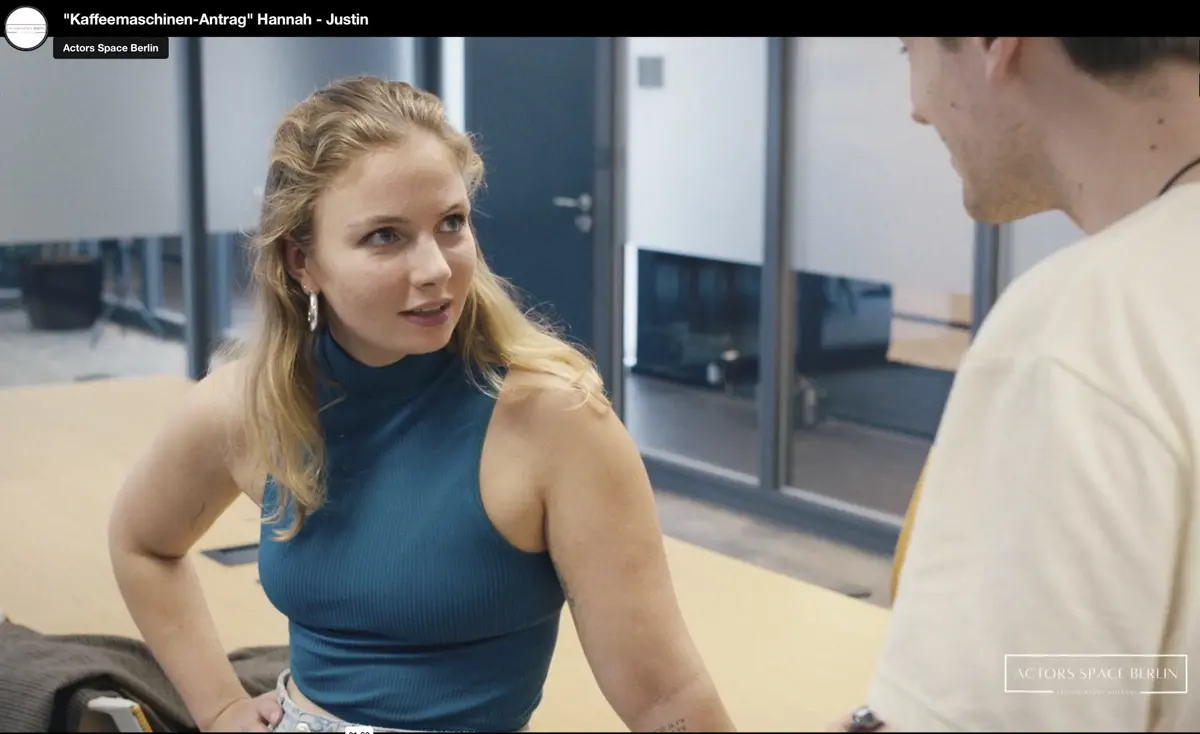 Eine Frau in einem ärmellosen blauen Oberteil steht an einem Schreibtisch und blickt aufmerksam zu einem Mann, der ihr gegenübersteht. Im Hintergrund sind Glastrennwände eines modernen Büros zu sehen.