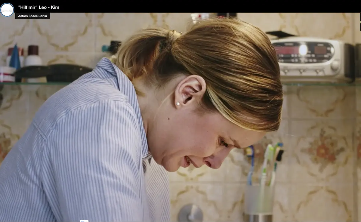 Eine Frau steht in einem Badezimmer mit floral gemusterten Fliesen und stützt sich über das Waschbecken, während sie emotional wirkt. Daneben stehen Zahnbürsten und Pflegeprodukte.