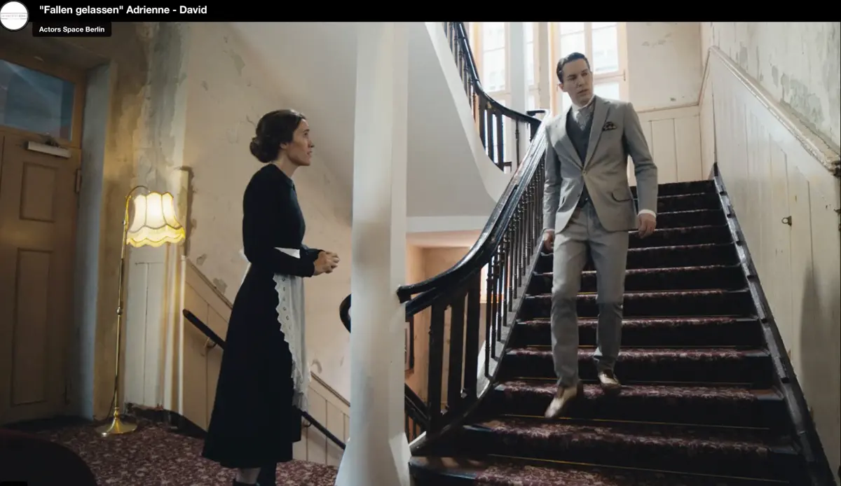 Eine Frau in historisch wirkender Dienstkleidung steht unten an einer Treppe und spricht mit einem Mann, der im Anzug ein paar Stufen höher steht. Die Szenerie spielt in einem älteren Gebäude mit Teppichboden und warmem Licht.