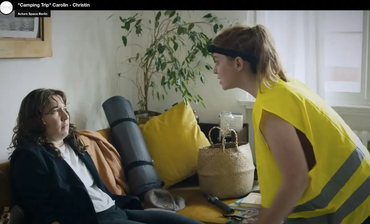 Eine Frau mit Trainingskleidung und gelber Warnweste lehnt sich nach vorn und spricht energisch mit einer anderen Frau, die erschöpft auf einem Sofa sitzt. Daneben stehen Campingausrüstung und Haushaltsgegenstände.