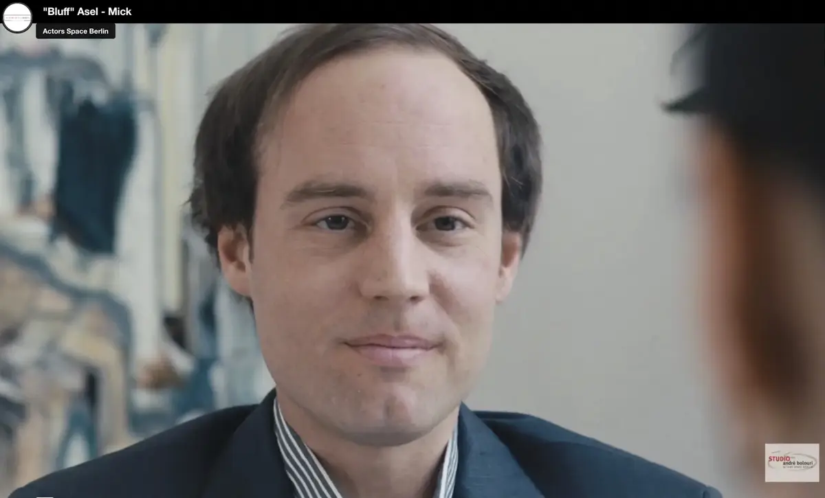 Nahaufnahme eines Mannes im Anzug, der lächelt und einer anderen Person gegenüber sitzt. Hinter ihm hängen abstrakte Kunstwerke.