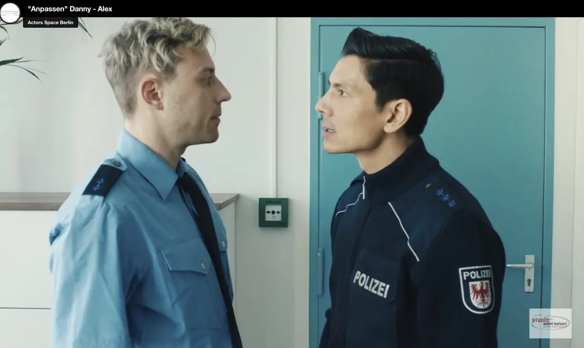 Zwei Männer in Polizeiuniformen stehen sich in einem hellen Raum gegenüber und diskutieren angespannt. Einer trägt ein hellblaues Hemd, der andere eine dunkle Einsatzjacke.