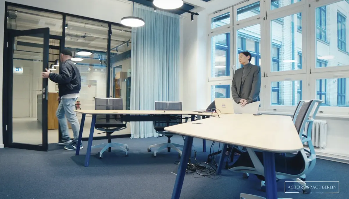 Ein Mann öffnet eine Tür und betritt einen modernen Besprechungsraum mit großen Fenstern und mehreren Schreibtischen. Eine zweite Person steht im Raum an einem Tisch mit Laptop.