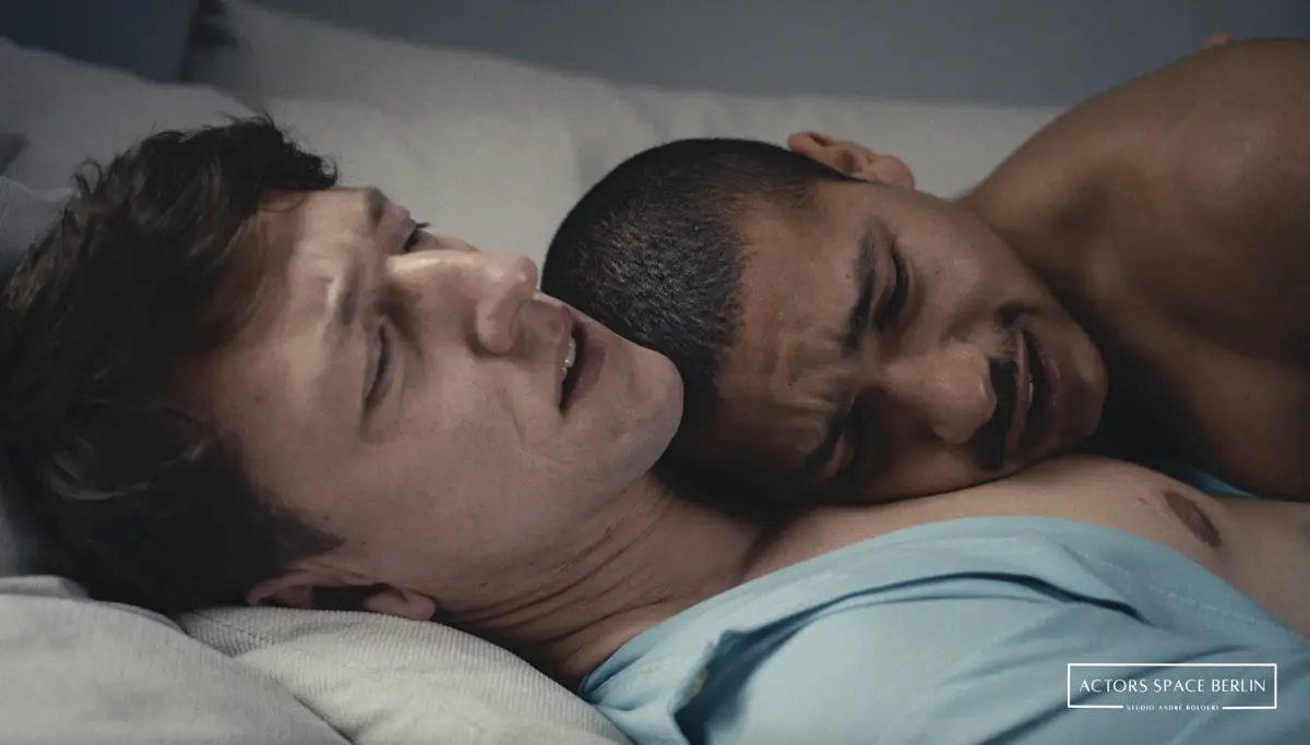 Zwei Männer liegen eng aneinandergeschmiegt im Bett. Einer lehnt halb auf dem Rücken, der andere liegt mit dem Kopf auf seiner Brust. Beide wirken emotional bewegt.