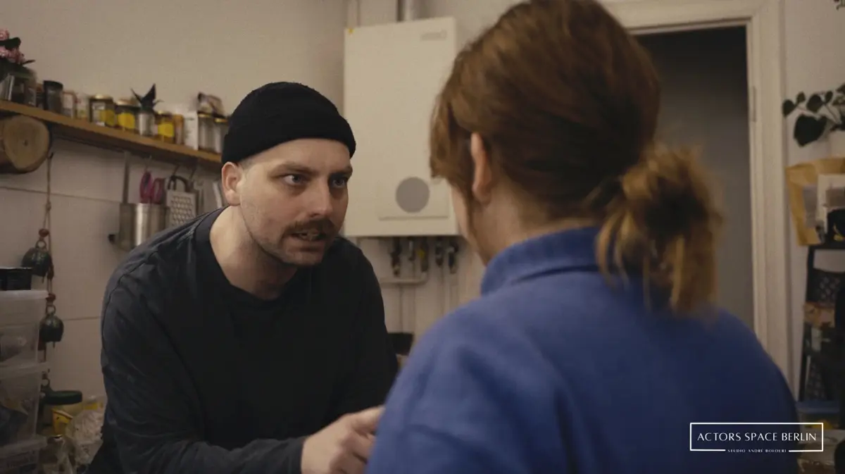 Ein Mann steht wütend oder aufgebracht an einem Konferenztisch, während eine blonde Frau ihm sitzend gegenüberschaut. Auf dem Tisch steht ein Laptop und eine Orchidee.