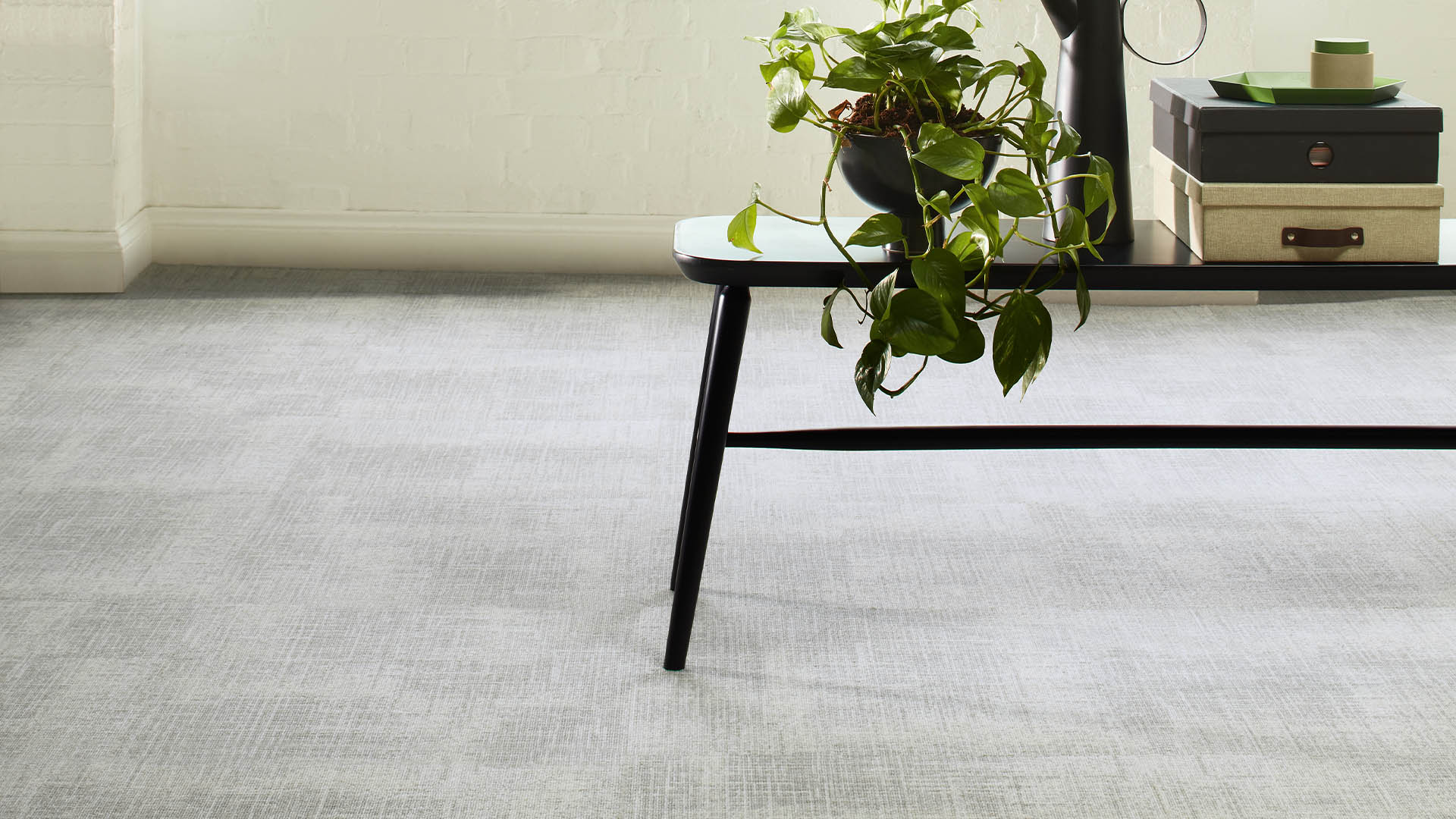 Neutral grey patterned carpet tiles in a bright office lobby with white brick walls, large industrial windows, and a minimalist black table with plants, demonstrating subtle textured flooring in a contemporary workspace.