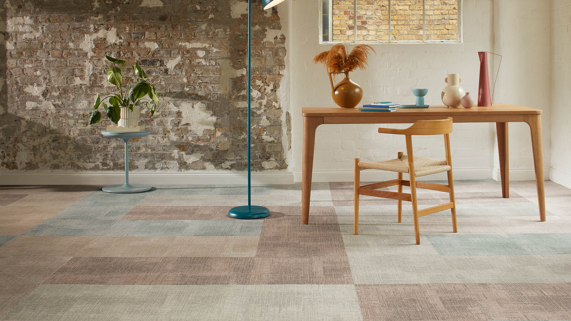 Patterned carpet tiles in clay, beige, and muted green earth tones in a contemporary office space with exposed brick wall, wooden desk, and modern furniture, showcasing warm biophilic-inspired flooring design.