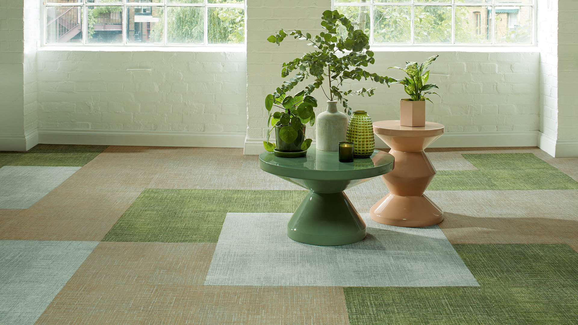 Green and neutral patterned carpet tiles in a modern office space with large industrial windows, indoor plants, and contemporary tables, highlighting biophilic-inspired flooring design.