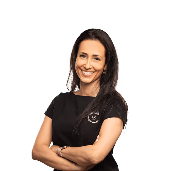 A smiling woman with long black hair, wearing a black shirt with a dental logo on it, standing with her arms crossed.