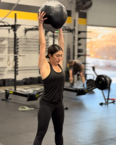 A woman in black athletic wear lifts a large medicine ball overhead at a gym. The space is equipped with fitness gear, and another person exercises in the background.