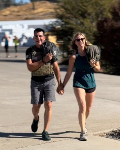 A joyful couple walks hand in hand on a sunny day, each carrying a backpack. The man smiles broadly; the woman grins, exuding happiness and warmth.