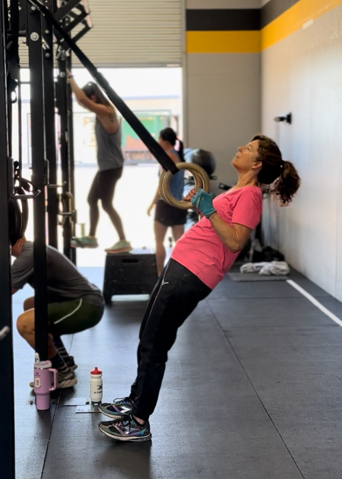 A woman in a pink shirt exercises on gymnastic rings in a gym, while others work out nearby. The scene conveys focus and determination.