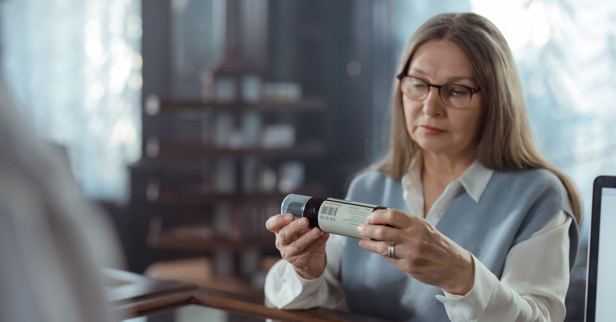 A senior woman with eyeglasses reviews a medicine bottle at a pharmacy counter indoors. — mark cuban pharmacy