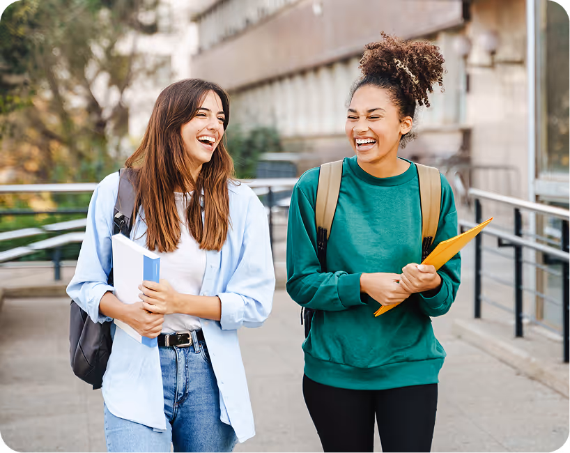 Two students laughing together on campus, carrying notebooks and backpacks