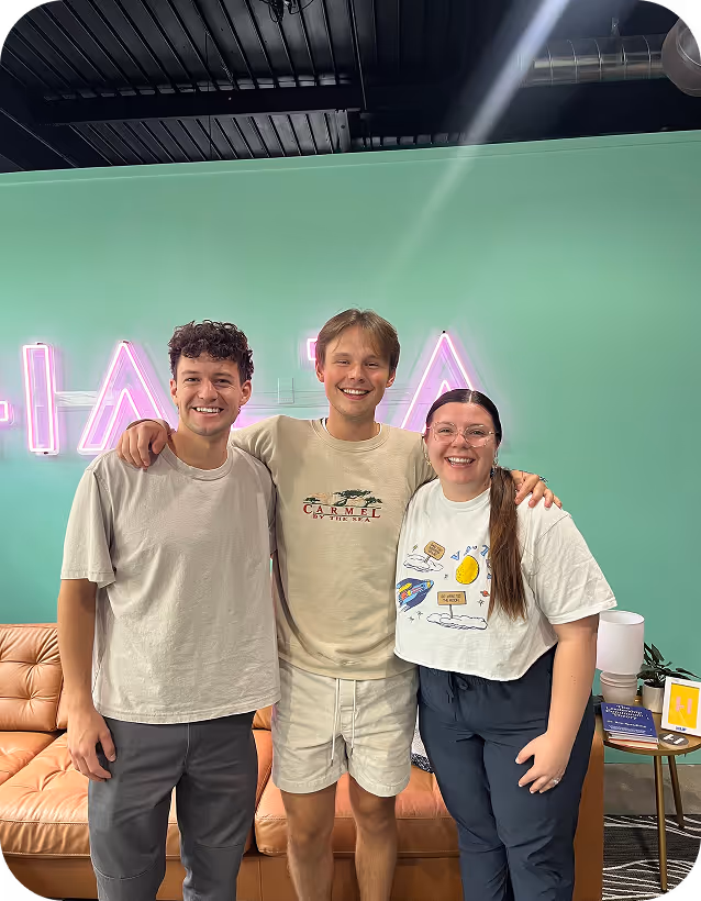 Three smiling friends posing together in front of green wall with neon sign