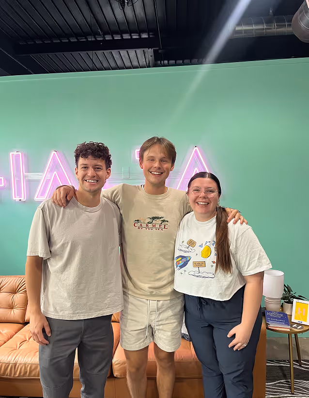 Three friends smiling together in front of green wall with neon sign