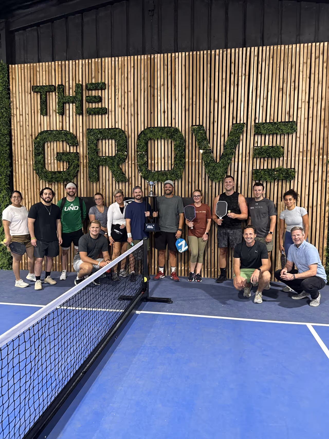 Group of people posing with pickleball paddles on an indoor court with "The Grove" sign