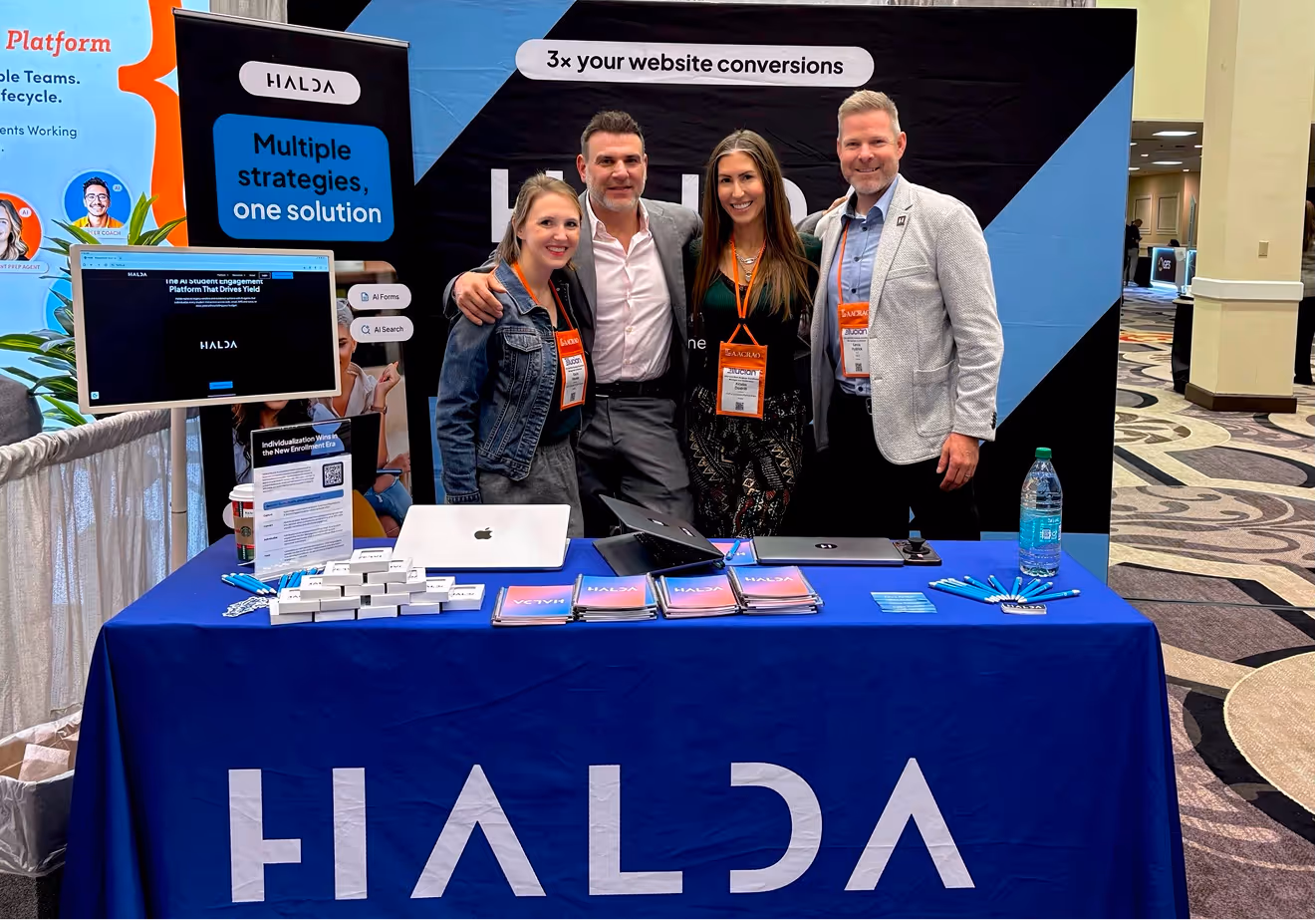 Four people with event badges smiling and posing behind a HALDA booth with laptops, brochures, and promotional items at a conference.
