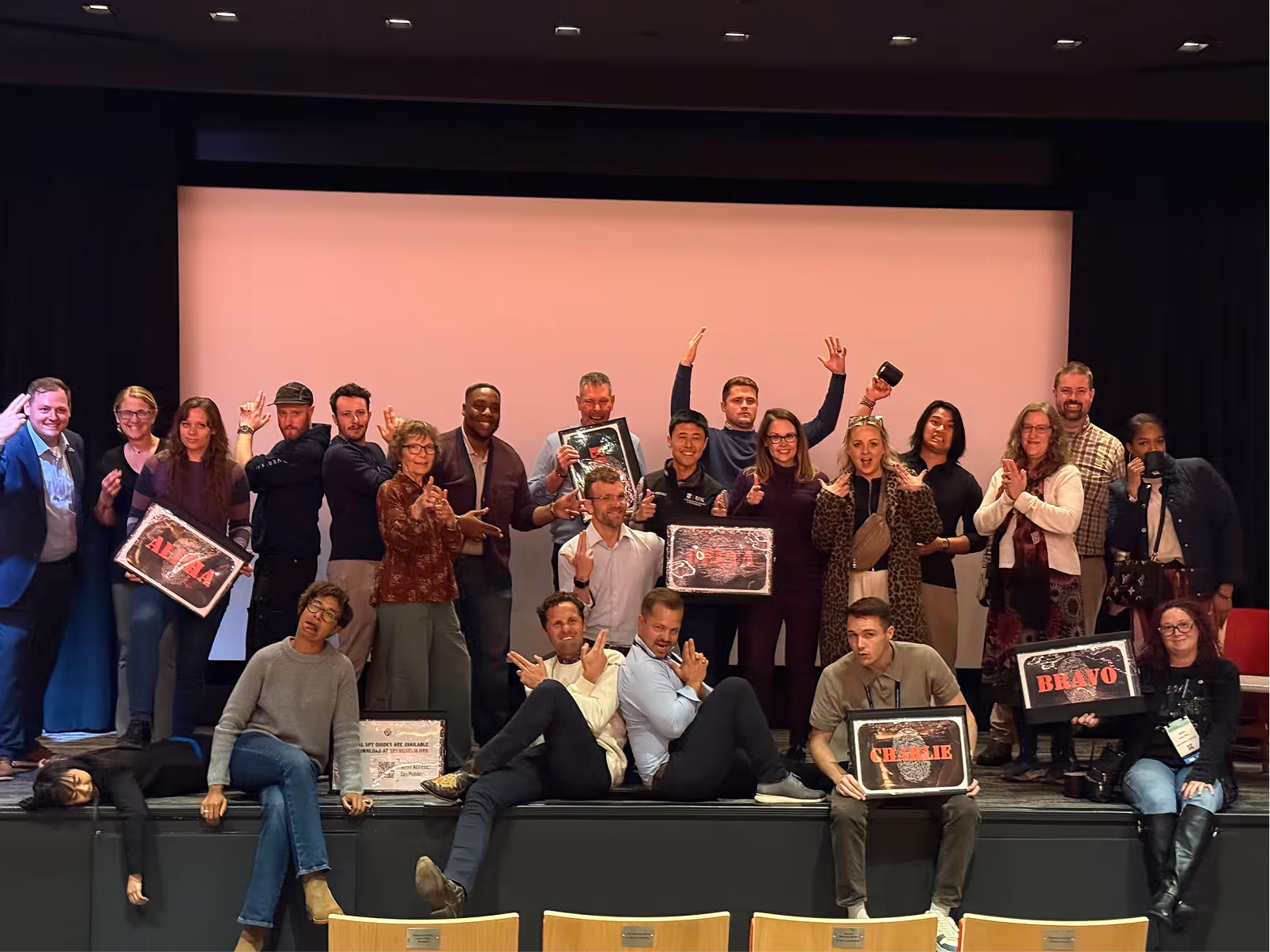 Group of people posing on a stage in front of a screen, some holding framed signs with names and striking playful hand gestures.