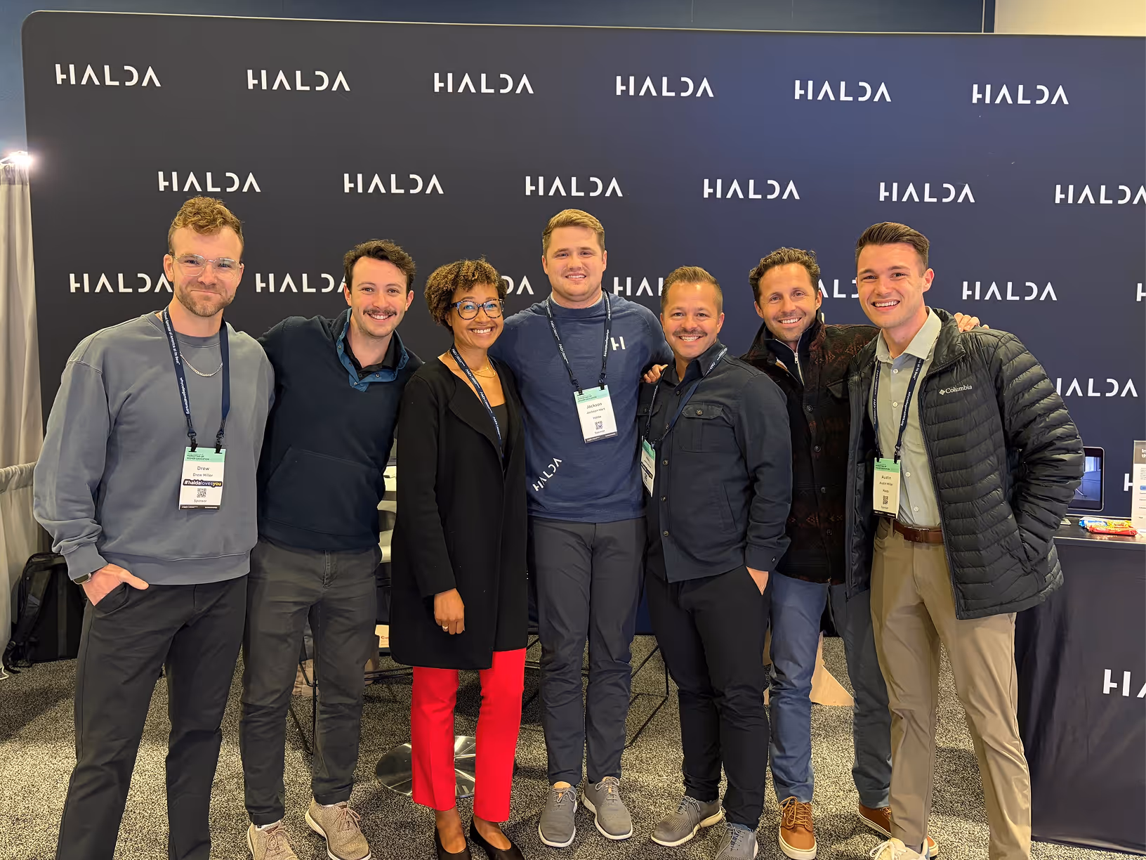 Group of seven people smiling and standing together in front of a black backdrop with multiple white HALDA logos.