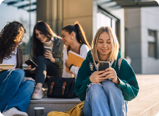 Four female students sitting on outdoor steps, three chatting while one in a green hoodie looks at her phone.