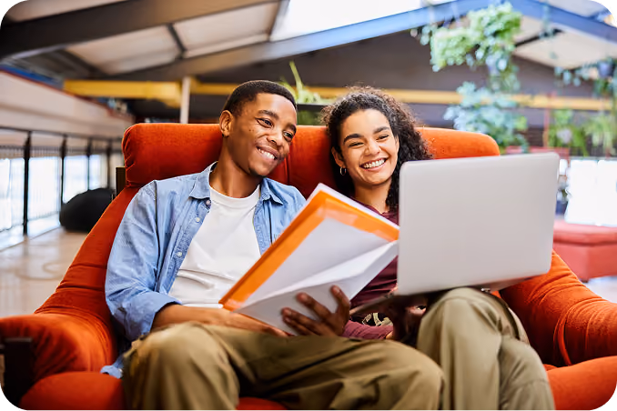 Two college students laughing together with a laptop and course materials in a campus lounge