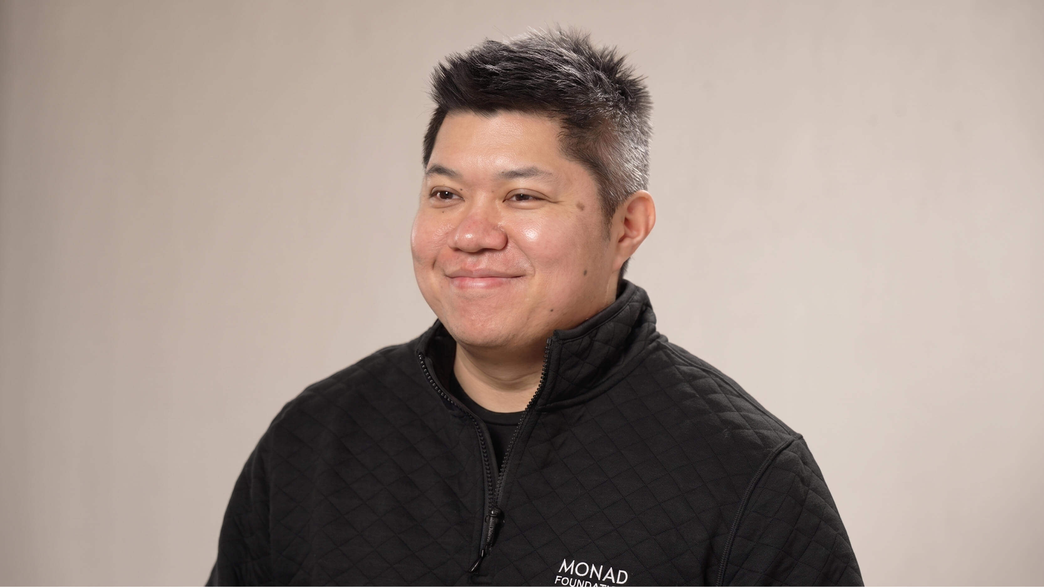 Smiling man with short black hair wearing a black Monad Foundation jacket against a plain background.