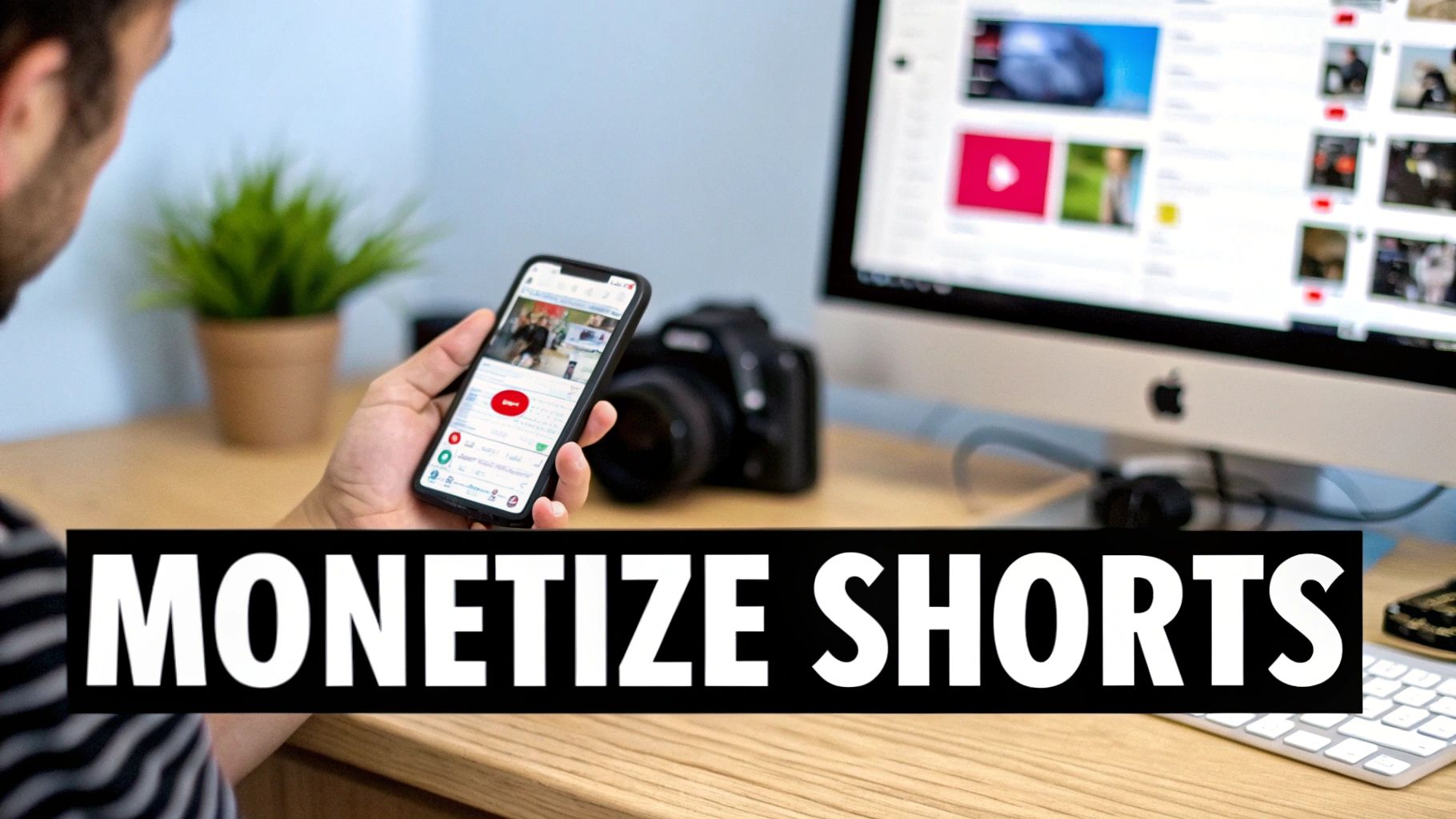 Person holding a smartphone displaying videos, with a computer monitor and camera on a wooden desk.