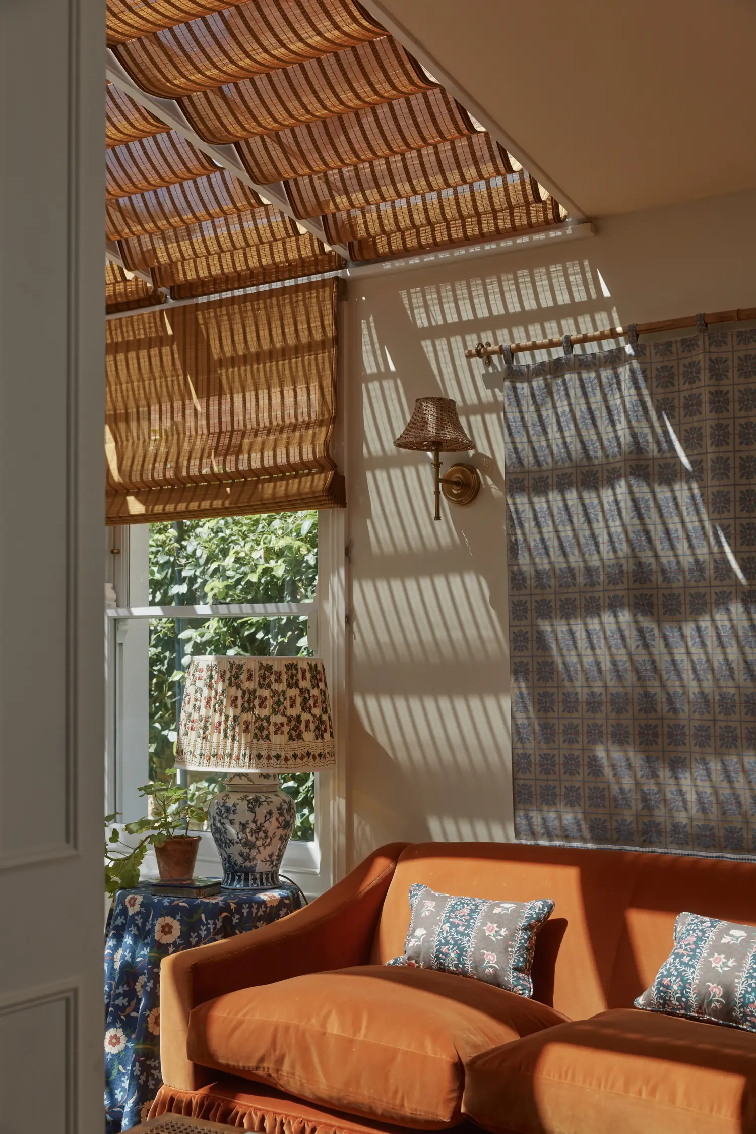 Bamboo Roman blind beside a patterned curtain and brass wall light, sunlight patterning the wall