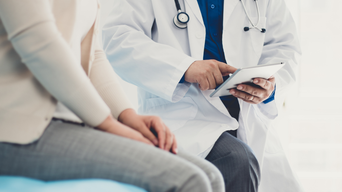 Doctor wearing a white coat and stethoscope holding a tablet while sitting next to a patient.