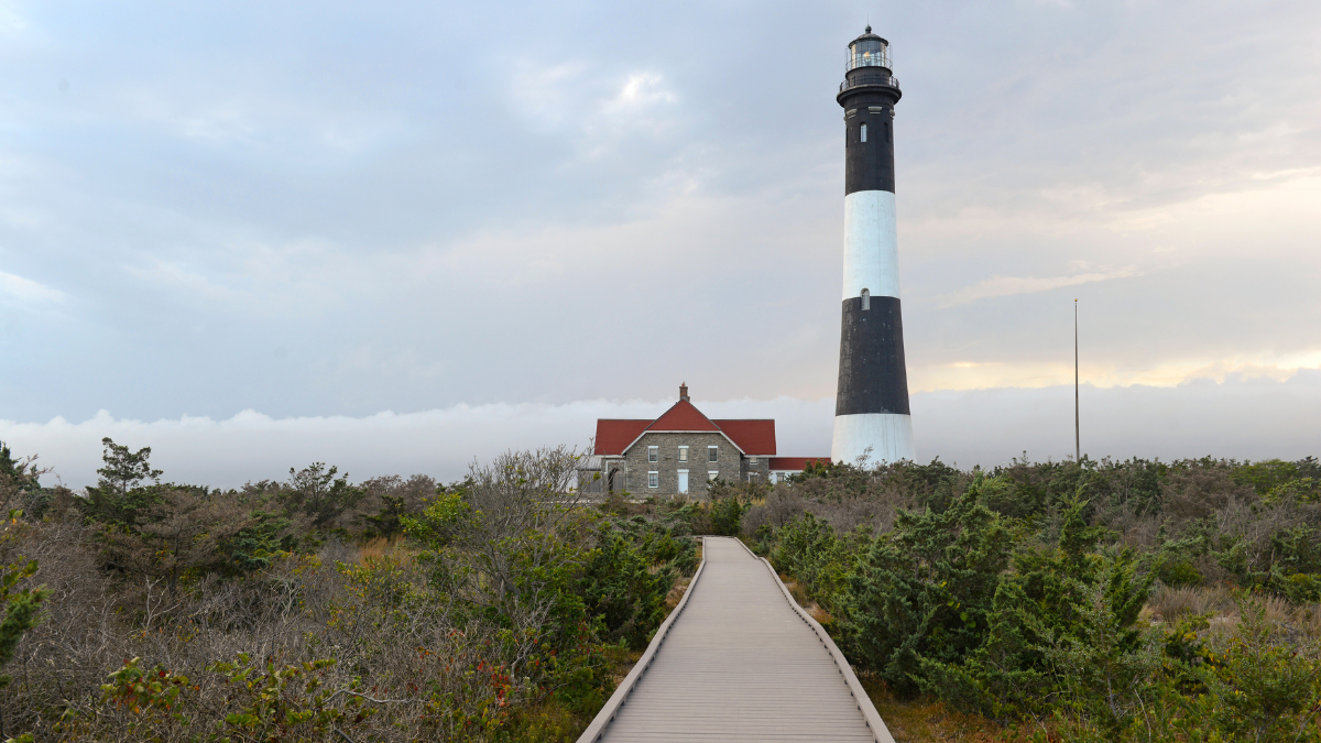 Black and white striped lighthouse in Long Island New York with red-roofed building along a wooden path surrounded by shrubs.
