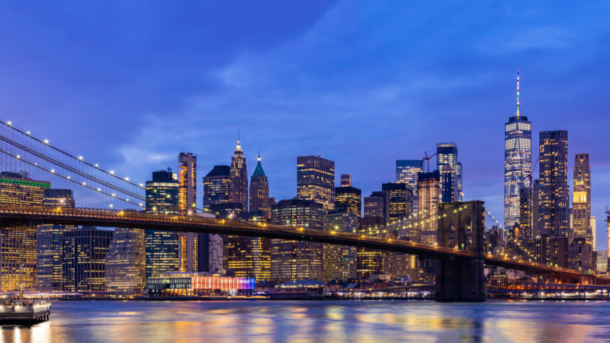 Brooklyn Bridge lit up at dusk with New York City skyline and One World Trade Center in the background.