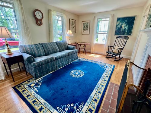 View of the living room standing next to the fireplace, showing 3 windows facing the lawn and neighbor’s house.