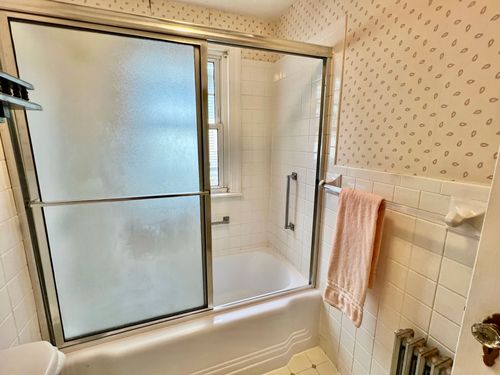 A bathroom with a sink, sliding door shower/bathtub, window, wood shelf, tile and wallpaper.