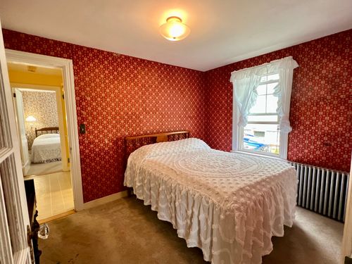A bedroom across the hall from the other bedroom, bathroom between them, with red wallpaper, glass door to the living room, window, and radiator.