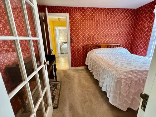 Same bedroom across the hall from the other bedroom, bathroom between them, with red wallpaper, glass door to the living room, window, and radiator.