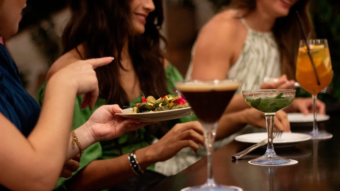 A view of a group of people passing tapas style plates to their left while drinking from visually aesthetic cocktails. 
