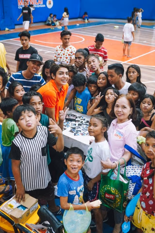 A group of children and two women smiling and posing around a table with photos in a gymnasium.