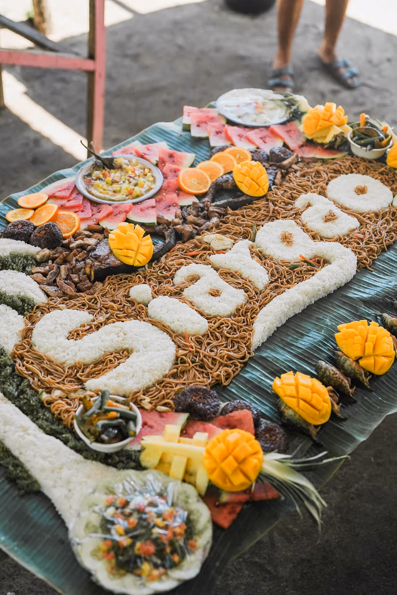 Food spread with rice spelling Siargao, surrounded by fruits, noodles, grilled meat, and dipping sauces.