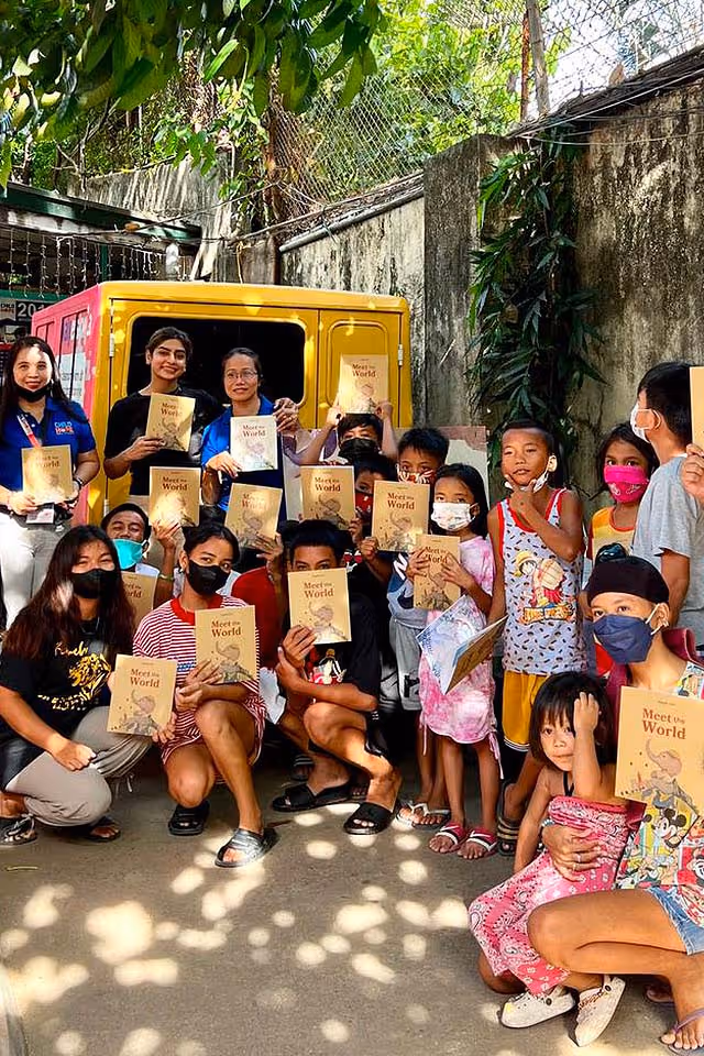 Group of adults and children outdoors holding 'Meet the World' booklets, some wearing face masks.