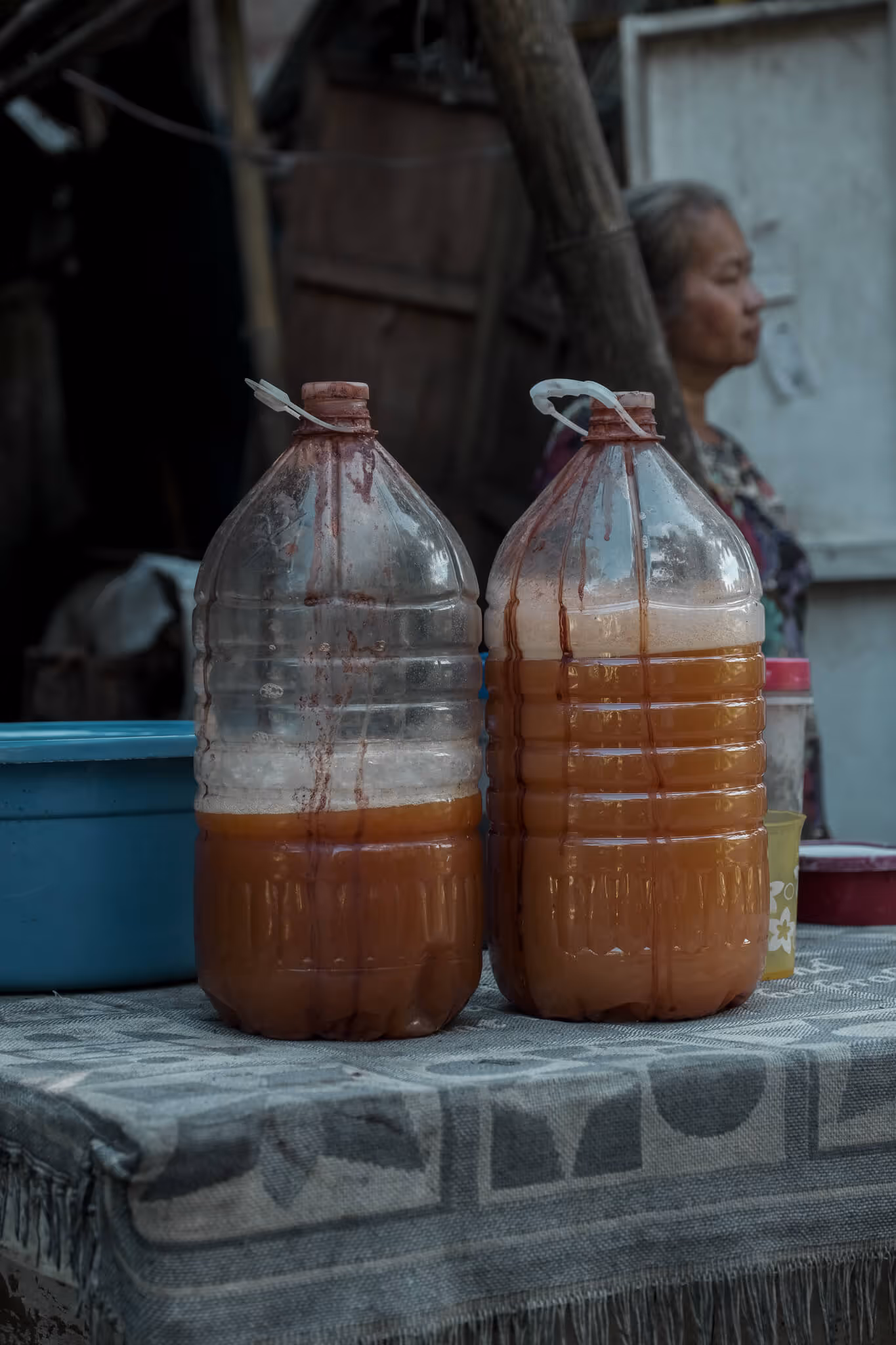 Two large plastic bottles with brownish liquid on a cloth-covered table, with a blurred person in background.