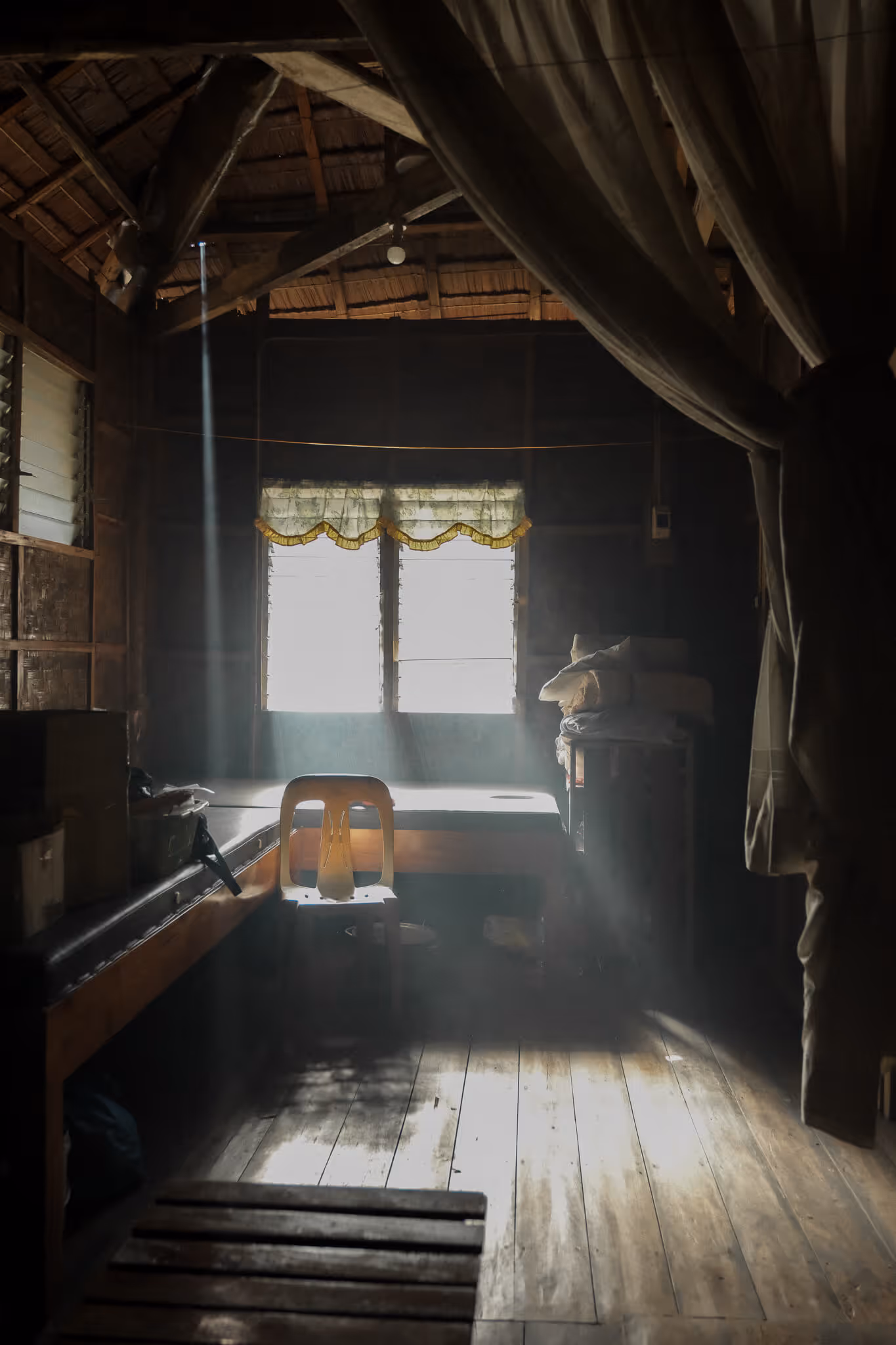 Sunlight streams through window onto a simple wooden room with a chair, bed, and stacked bedding.