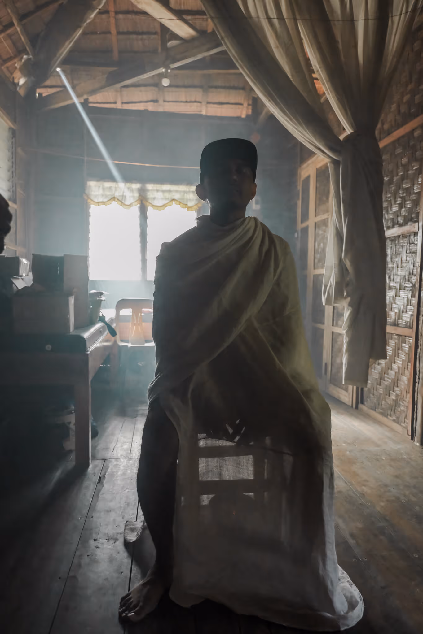 Silhouetted person sitting on a chair wrapped in cloth inside a rustic wooden room with light beams.