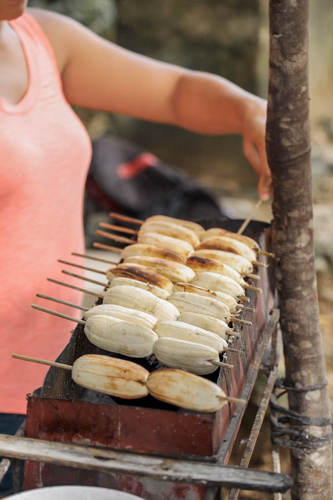 Woman grilling skewered bananas on a rectangular charcoal grill outdoors.