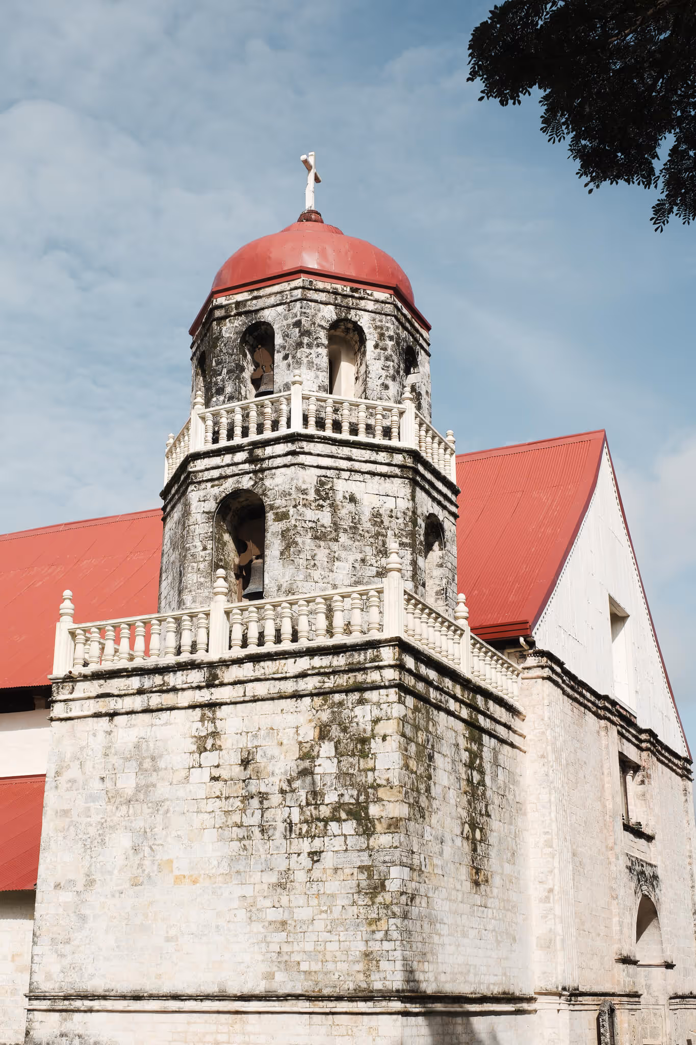 Stone church bell tower with red dome roof and a white cross on top under a blue sky.