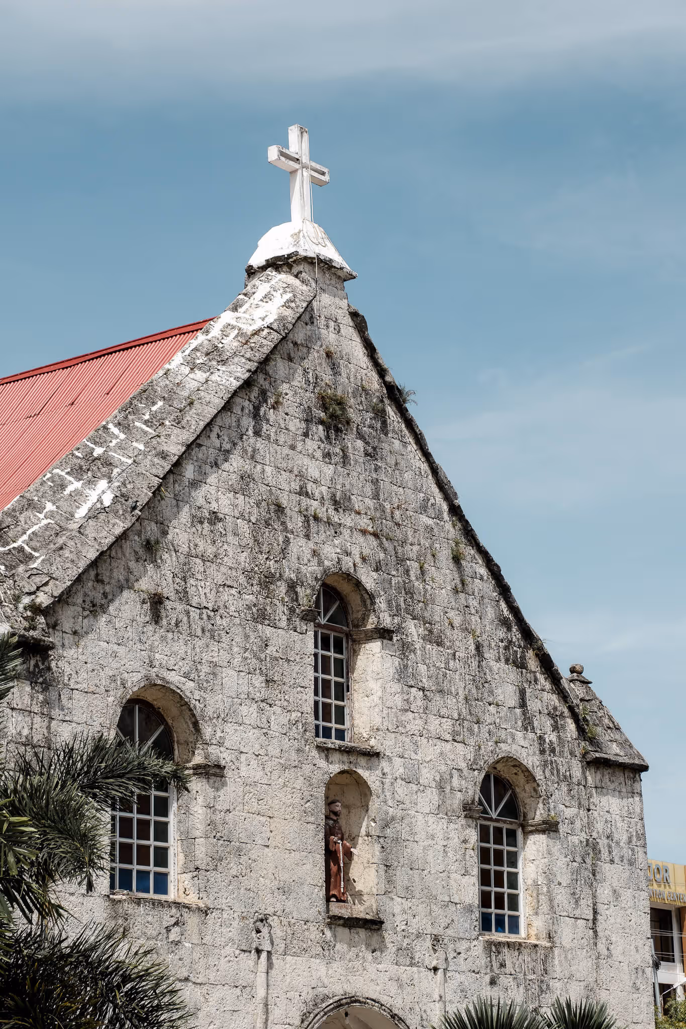 Stone church facade with arched windows, a statue in a niche, red roof, and a white cross on top.